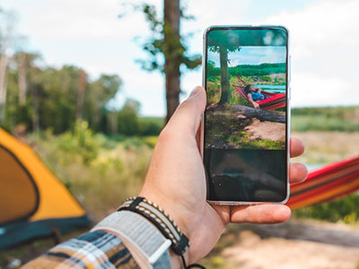 A person holds a smartphone, capturing a camping scene with a hammock and yellow tent near a lake. 