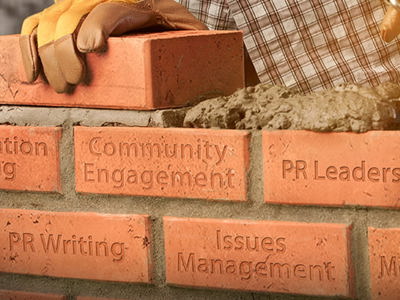 A close-up of a brick wall being built, with a gloved hand placing a brick and a trowel adding mortar. Each brick is etched with a public relations concept: Communication Planning, Community Engagement, PR Leadership, PR Writing, Issues Management, and Media Relations. This image visually represents building foundational skills for a successful PR career.
