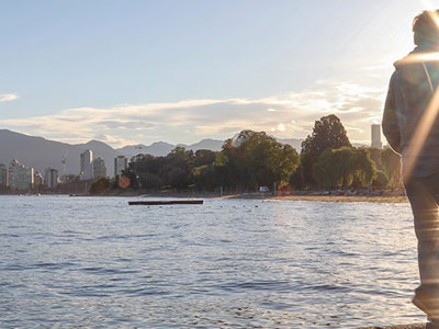Person walking along a body of water with an urban skyline on the other side of the water.